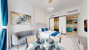 Une chambre d’hôtel moderne avec un canapé, un lit, un coin repas avec une table dressée pour deux personnes, et une cuisine en arrière-plan.