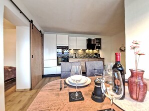 Uma área de refeições com mesa de madeira posta para dois, uma garrafa de vinho e um vaso com flores.