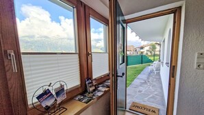 Ein Zimmer mit Fensterblick auf einen Garten, eine Tür mit Fußmatte und ein Fensterbrett mit Zeitschriften und einer kleinen Pflanze.