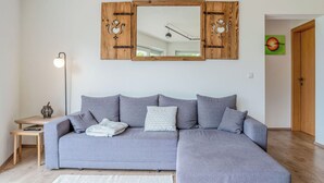 Un sofá seccional gris con una manta blanca y almohadas estampadas, una pequeña mesita de madera con una lámpara, y una alfombra blanca sobre piso de madera.