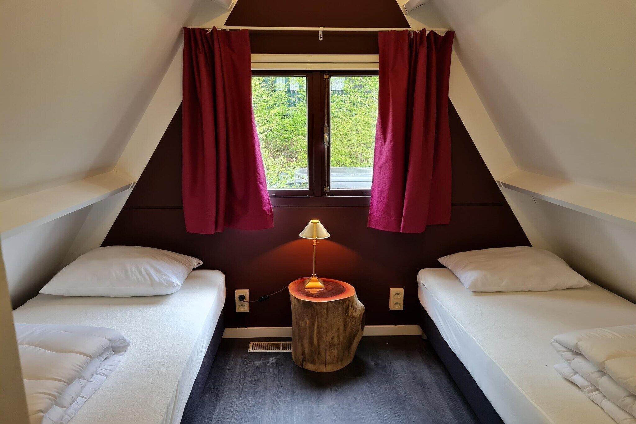 A small, triangular room with two beds, a wooden nightstand with a lamp, and a window with curtains.