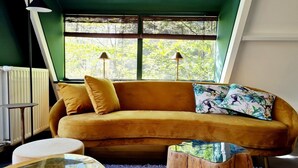 Una habitación con un sofá color mostaza, una mesa de centro redonda de madera y una ventana con persianas.