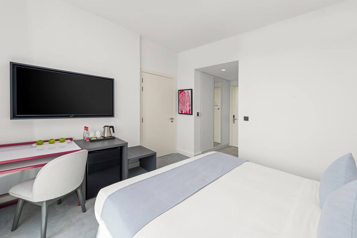 Una habitación de hotel moderna con cama, escritorio con televisor de pantalla plana, silla y pequeña área de kitchenette.