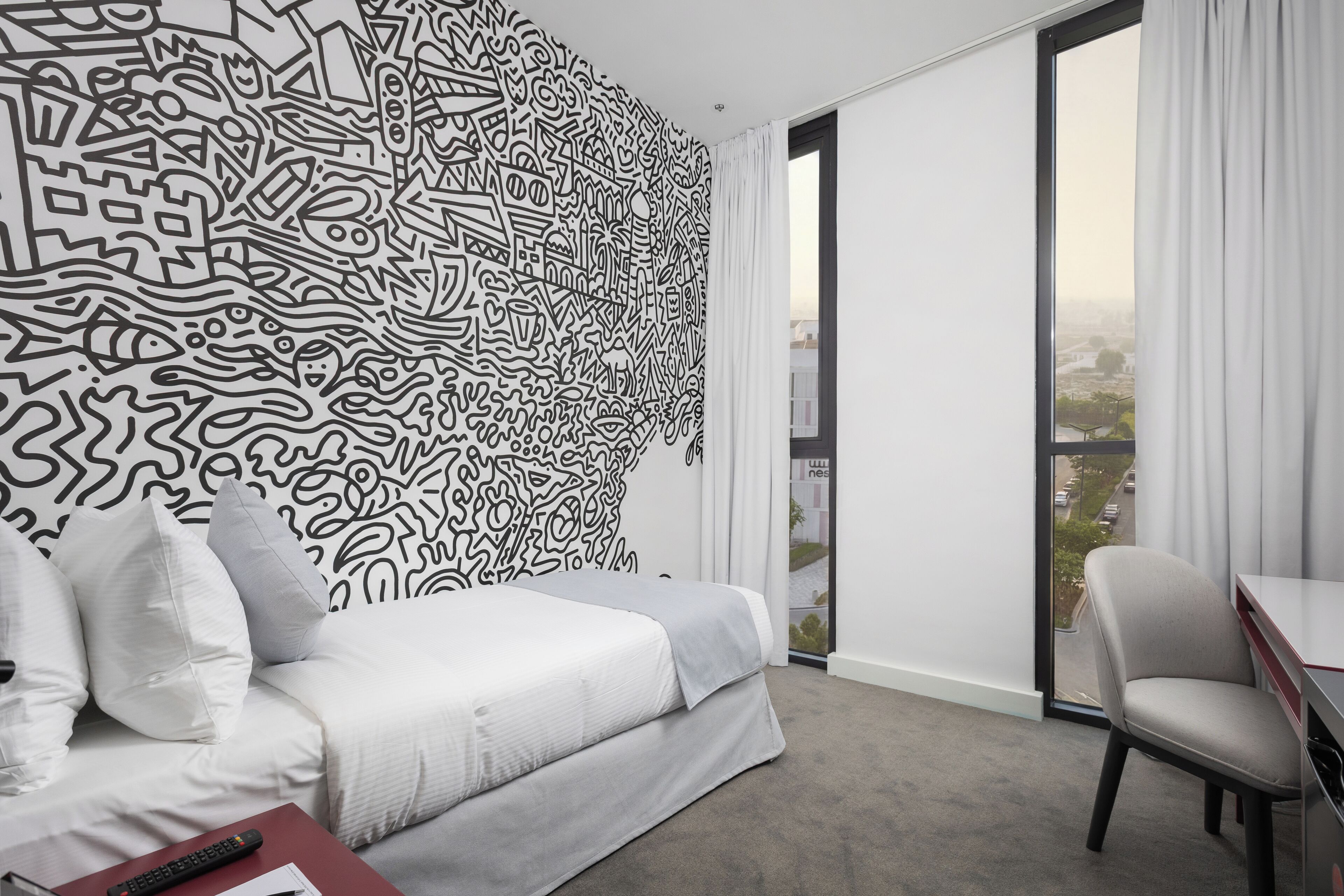 Een moderne hotelkamer met een groot abstracte muurschildering, een bed met wit beddengoed, een grijze bank, een bureau met een stoel, en een uitzicht op een stadsgezicht door een groot raam.