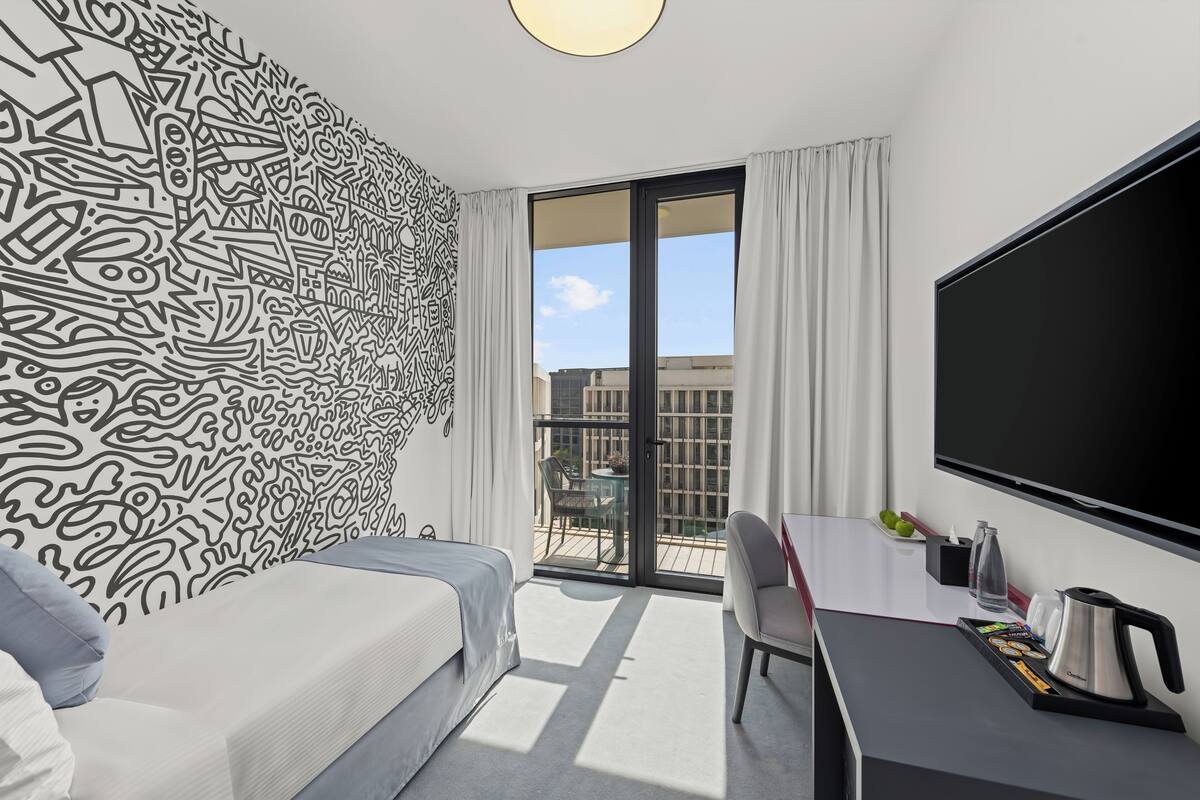 Habitación de hotel moderna con una cama grande, un escritorio con hervidor, un televisor de pantalla plana y un balcón con vistas a edificios.