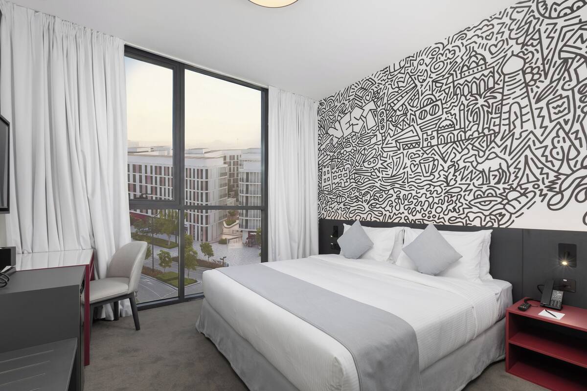 Habitación de hotel moderna con una cama grande, un escritorio y una silla. La habitación tiene un ventanal con vistas a edificios.