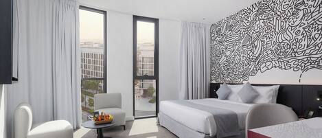 A modern hotel room with a large bed, a sitting area with a table and chair, a small balcony, and a decorative wall.
