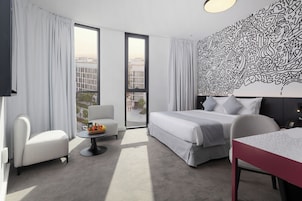 Una habitación de hotel moderna con una cama grande, un rincón de estar con una mesa y una silla, un balcón pequeño y un adorno en la pared.