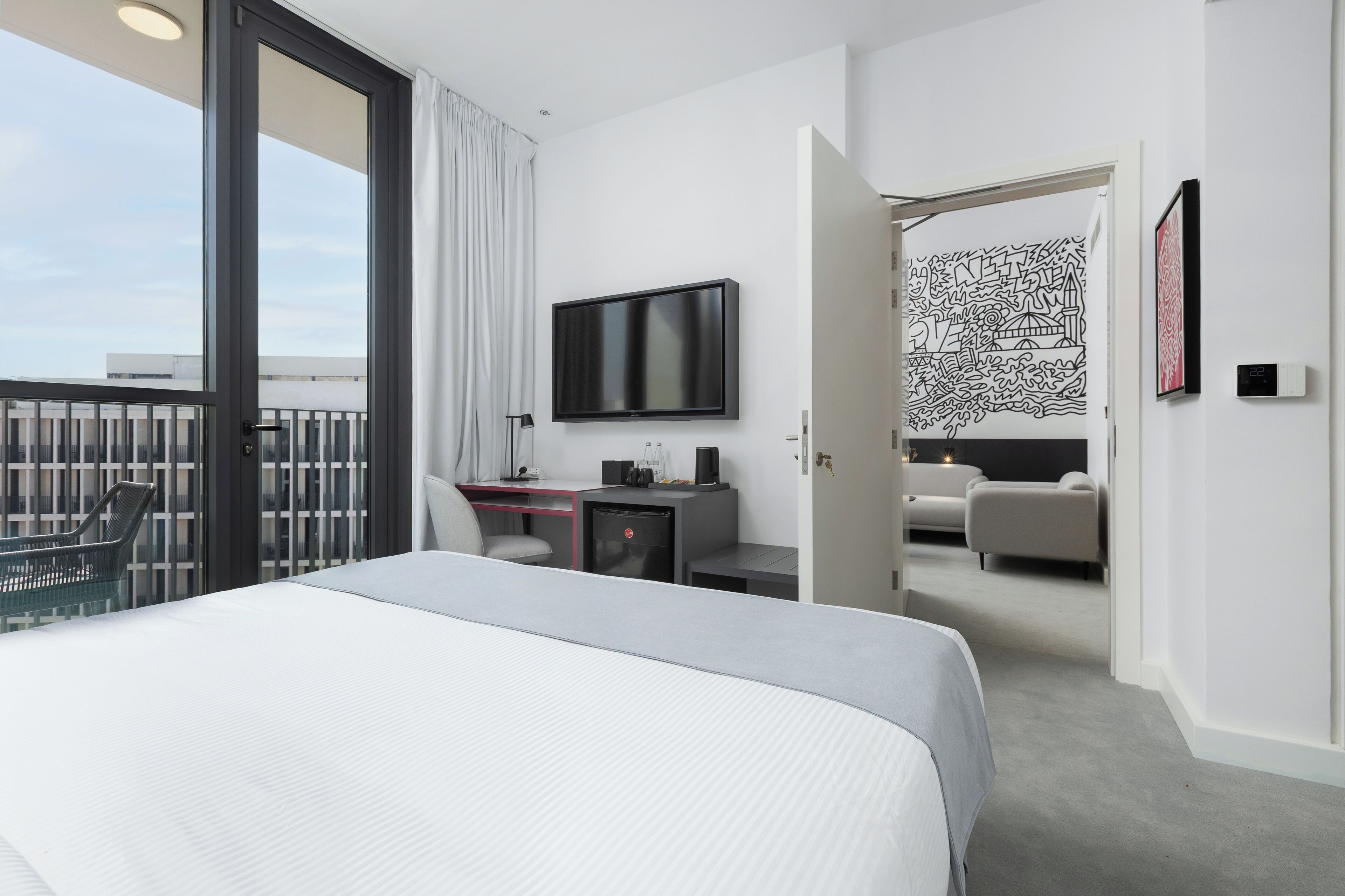 A modern hotel room with a large bed, a desk with a flat-screen TV, a small refrigerator, and a balcony with a view of the city.