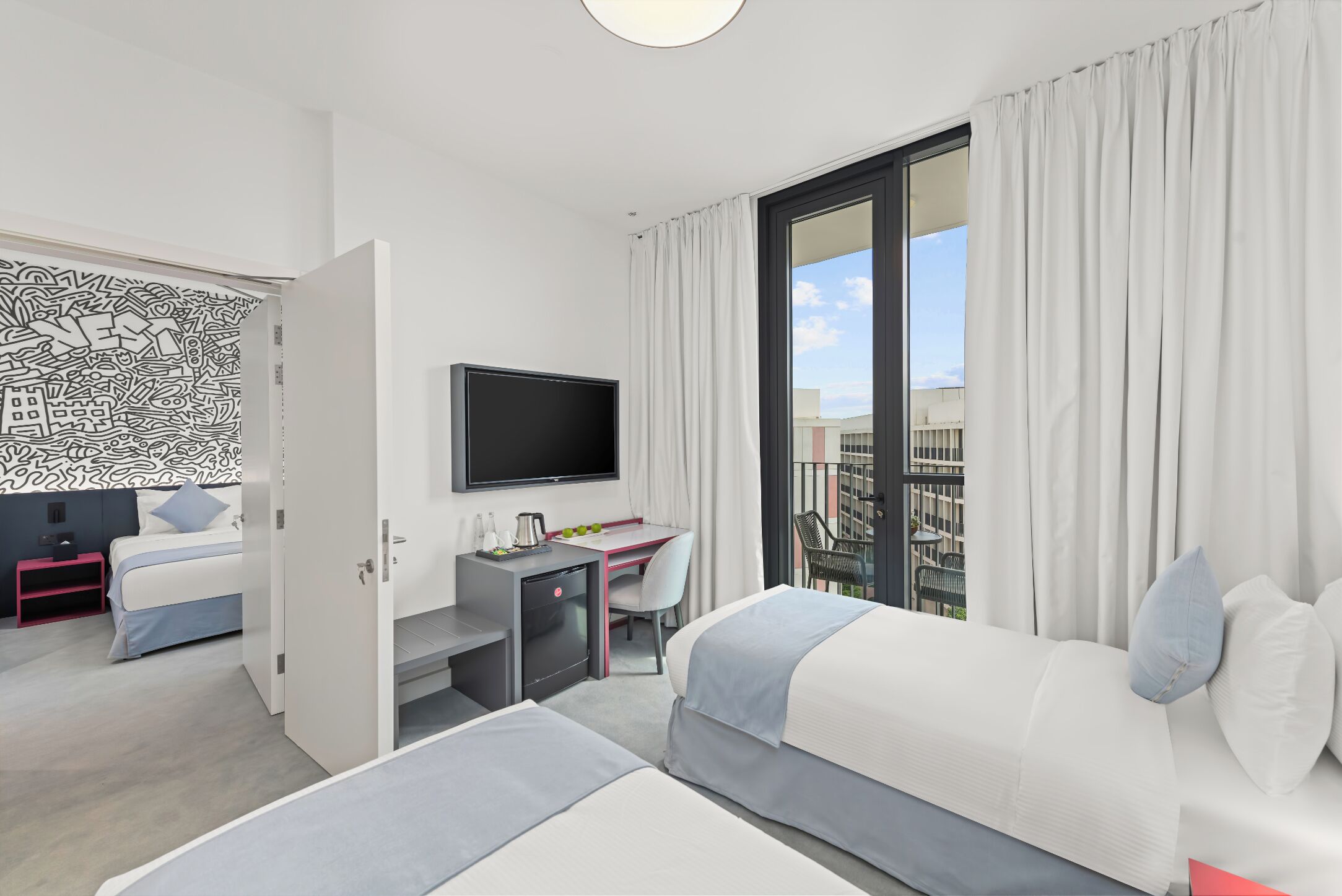 A modern hotel room with a bed, a desk with a TV, a balcony with curtains, and a wall with abstract art.
