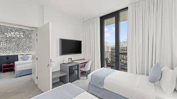 A modern hotel room with a bed, a desk with a TV, a balcony with curtains, and a wall with abstract art.