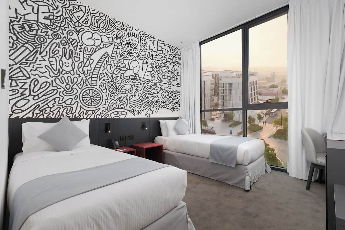 Habitación de hotel con dos camas, un ventanal con vistas a edificios y una pared con un diseño abstracto en blanco y negro.