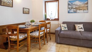 A dining area with a wooden table set for four, flanked by chairs with floral cushions. A gray sofa with patterned cushions is against the wall, and a window with curtains is visible. - Lovely apartment for 4 people with WIFI, TV and pets allowed (Isolaccia)