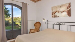 A bedroom with a bed, a chair, a lamp, a window with curtains, and a painting on the wall.