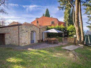 Un bâtiment en pierre avec un toit en tuiles, une porte en bois et un patio comprenant une table et des chaises, le tout abrité par un parasol blanc.