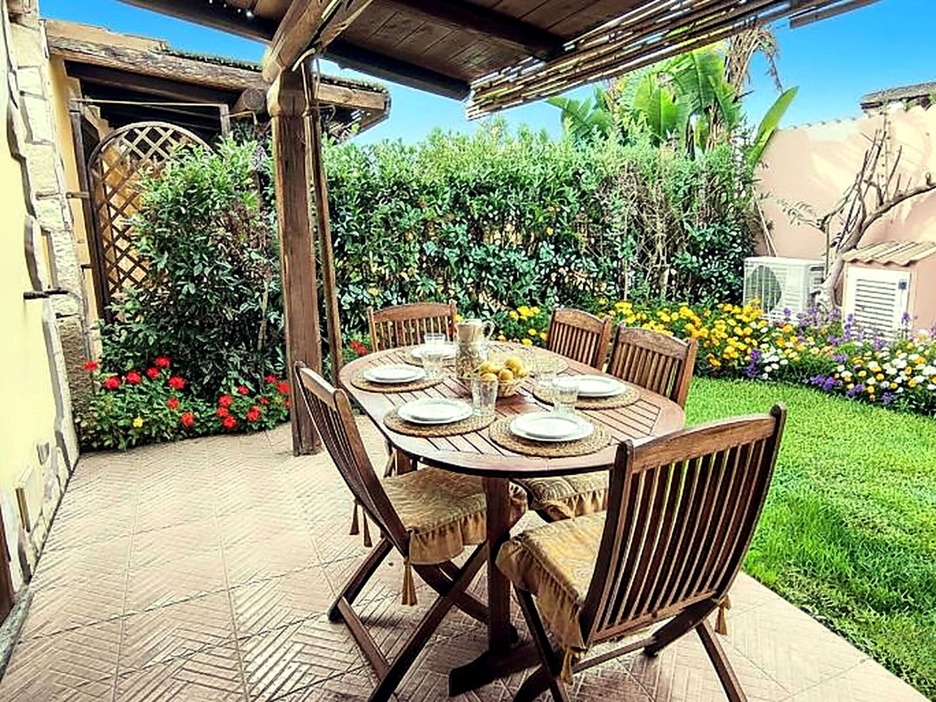 An outdoor patio with a wooden table set for four, surrounded by chairs, in a garden with various plants and flowers.