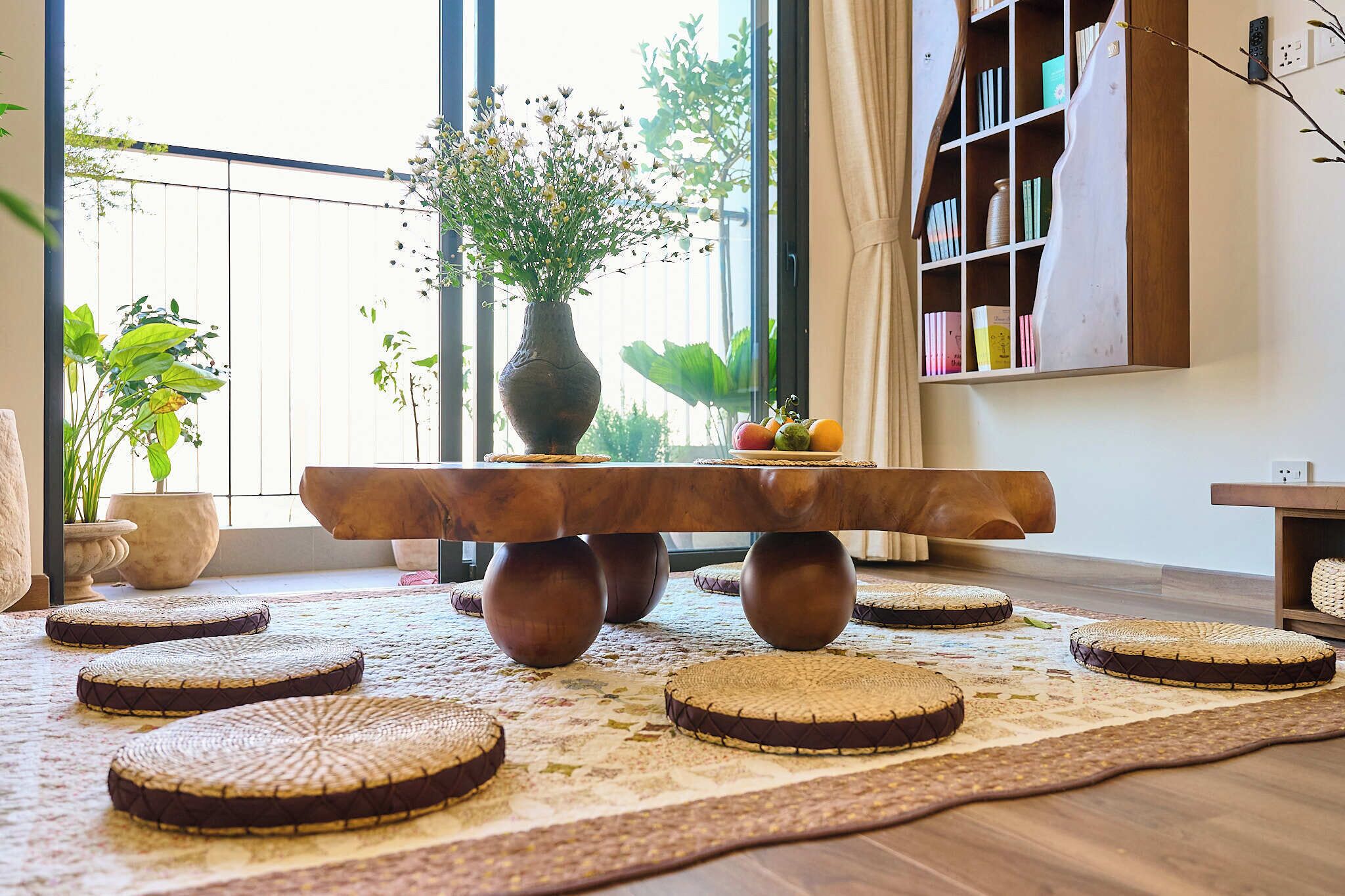 Een houten tafel met een bloemenvaas en fruit, omringd door geweven krukken op een vloerkleed met patroon in een lichte kamer met grote ramen en een boekenkast.
