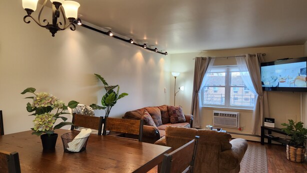 A cozy living room with a wooden dining table, comfortable seating, a TV, and a large window with curtains.