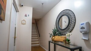 A hallway with a wooden floor, a black-framed mirror, a hand sanitizer dispenser, a welcome sign, and a small plant on a console table.