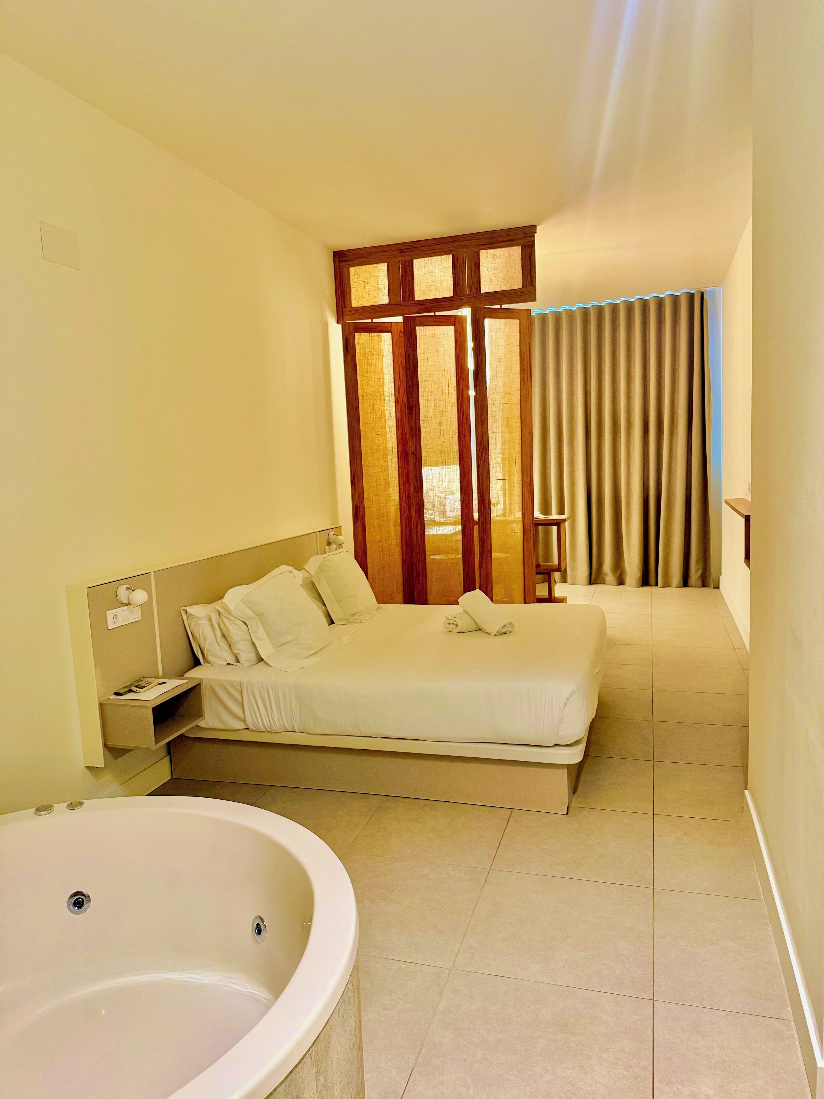 A hotel room with a large bed, a bedside table with a lamp, a bathroom with a bathtub, and a view of the outside through a window with curtains.