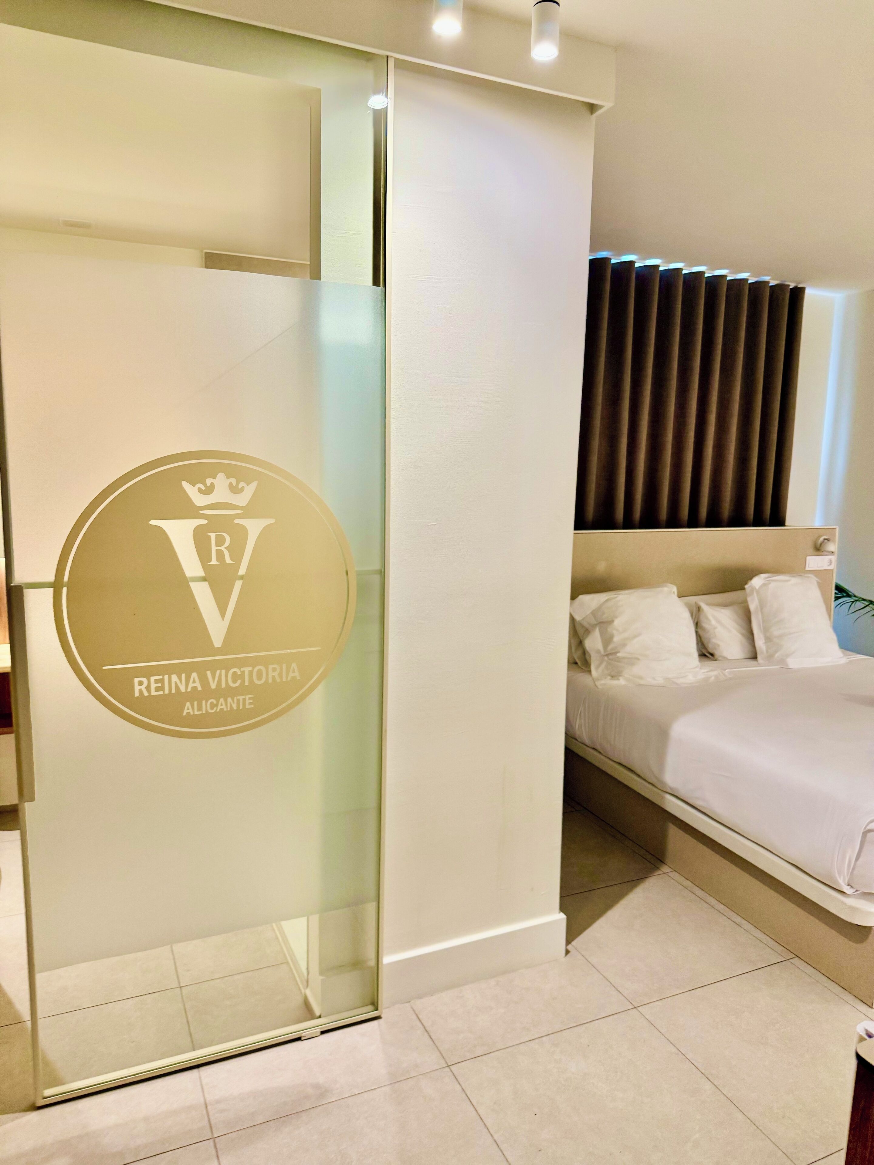 Una habitación de hotel moderna con cama, un separador de vidrio con un logotipo y una ventana cubierta con cortinas.