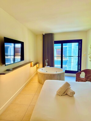 Ein Hotelzimmer mit einem großen Bett, einer runden Badewanne, einem an der Wand montierten Fernseher und einem Balkon mit Blick.