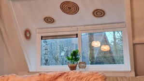 A room with a sloped ceiling, a large window with a view of trees, a small plant, and a wicker chair. - Renovated Ardennes Chalet with Patio (Durbuy)