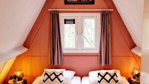A cozy, triangular room with two beds, a window with curtains, and a painting on the wall. - Renovated Ardennes Chalet with Patio (Durbuy)