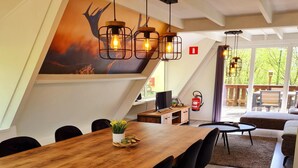 A dining area with a wooden table, black chairs, and pendant lighting. There is a painting on the wall and a view of the outdoors through large windows. - Comfortable Fenced Chalet in Durbuy (Durbuy)