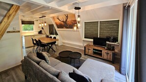 A modern loft-style living space with a dining area, a large sofa, a TV, and a painting of a deer. - Comfortable Fenced Chalet in Durbuy (Durbuy)