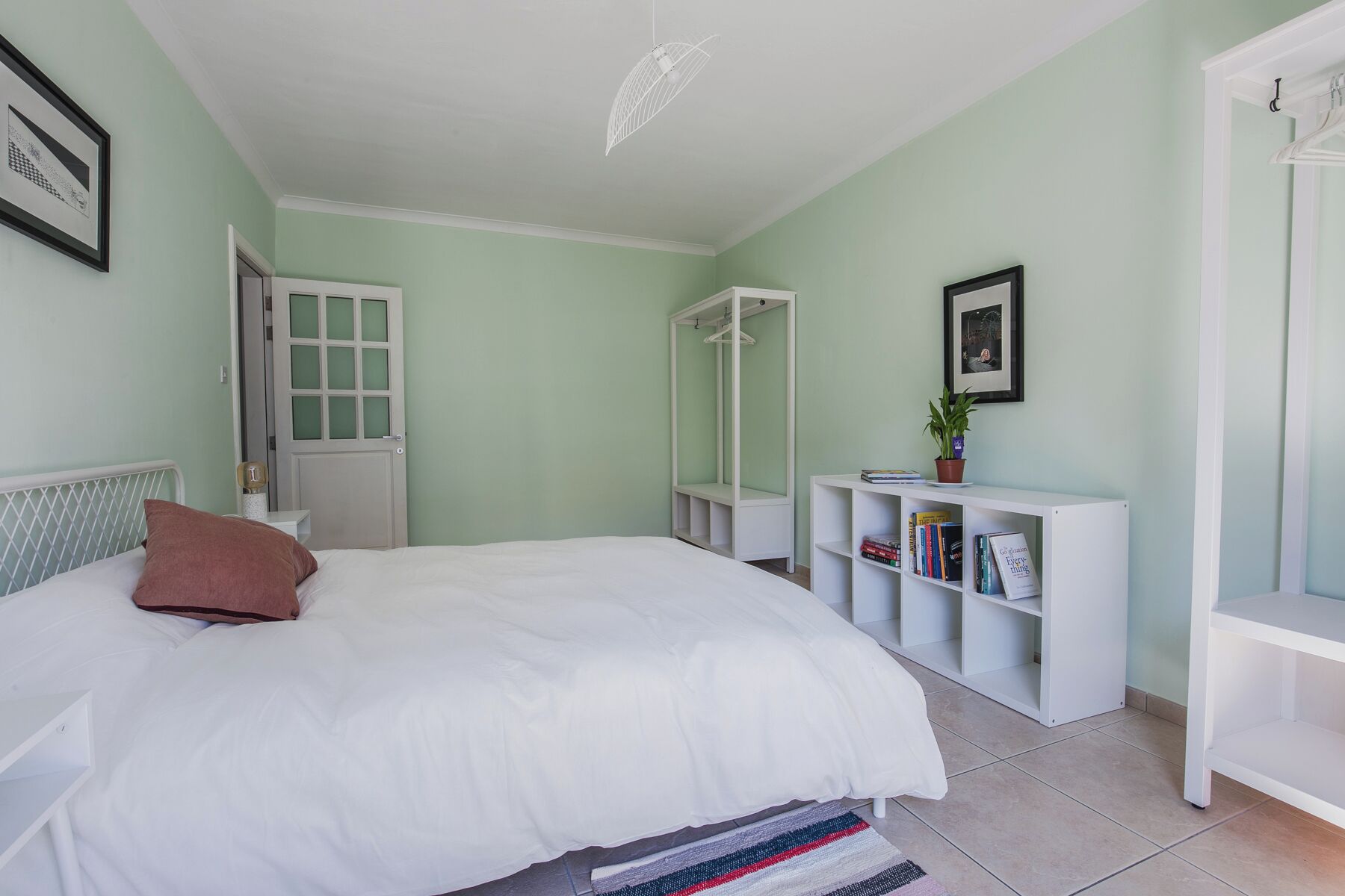 A bedroom with a bed, a wardrobe, a mirror, and a shelf with books and a plant.