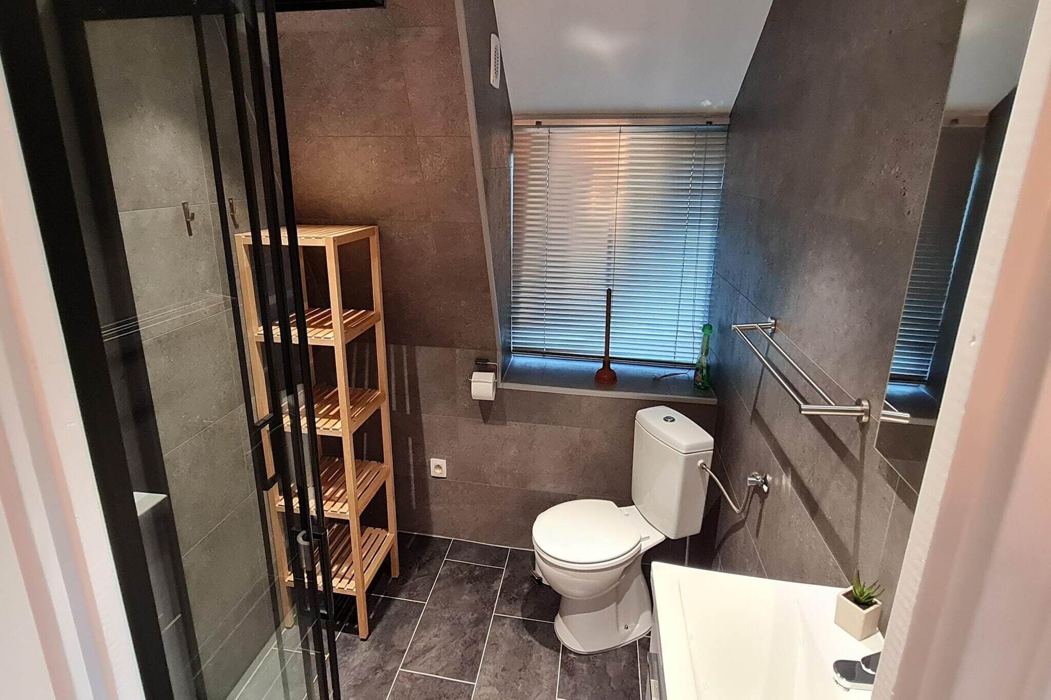 Een compacte badkamer met toilet, een klein raam met zonwering, een houten plank en een glazen deur.