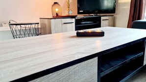 Ein modernes Wohnzimmer mit Couchtisch aus Holz, einem Fernseher auf einem Schrank, einem kleinen Beistelltisch mit Lampe und einem Sessel.