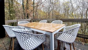 Eine Holzterrasse mit Tisch und weißen Gitterstuhlen, umgeben von Bäumen.