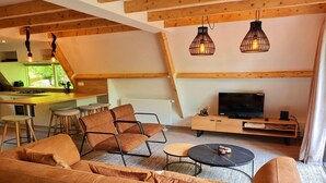 Ein modernes Wohnzimmer mit einer Couch, einem Couchtisch, Sesseln, einer Küchenzeile mit Barhocker und einem Fernseher auf einer Holzkommode.