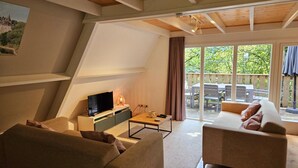 Uma sala de estar moderna com um sofá, uma mesa de centro e uma televisão. Há uma janela grande com cortinas, e uma varanda com móveis de exterior.