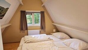 Quarto com teto inclinado, janela com cortinas, radiador e duas camas com roupa de cama branca.