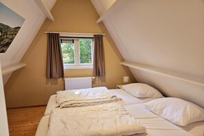Ein Schlafzimmer mit schrägen Wänden, einem fenster mit Vorhängen, einer Heizung und zwei Betten mit weißer Bettwäsche.