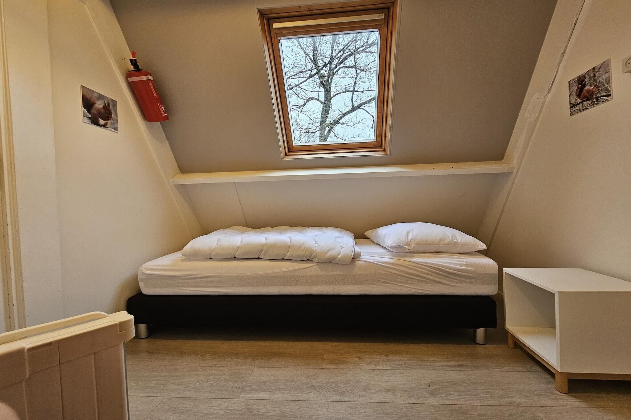 Een slaapkamer met een laag schuin dak, een enkel bed, een raam en een brandblusser.