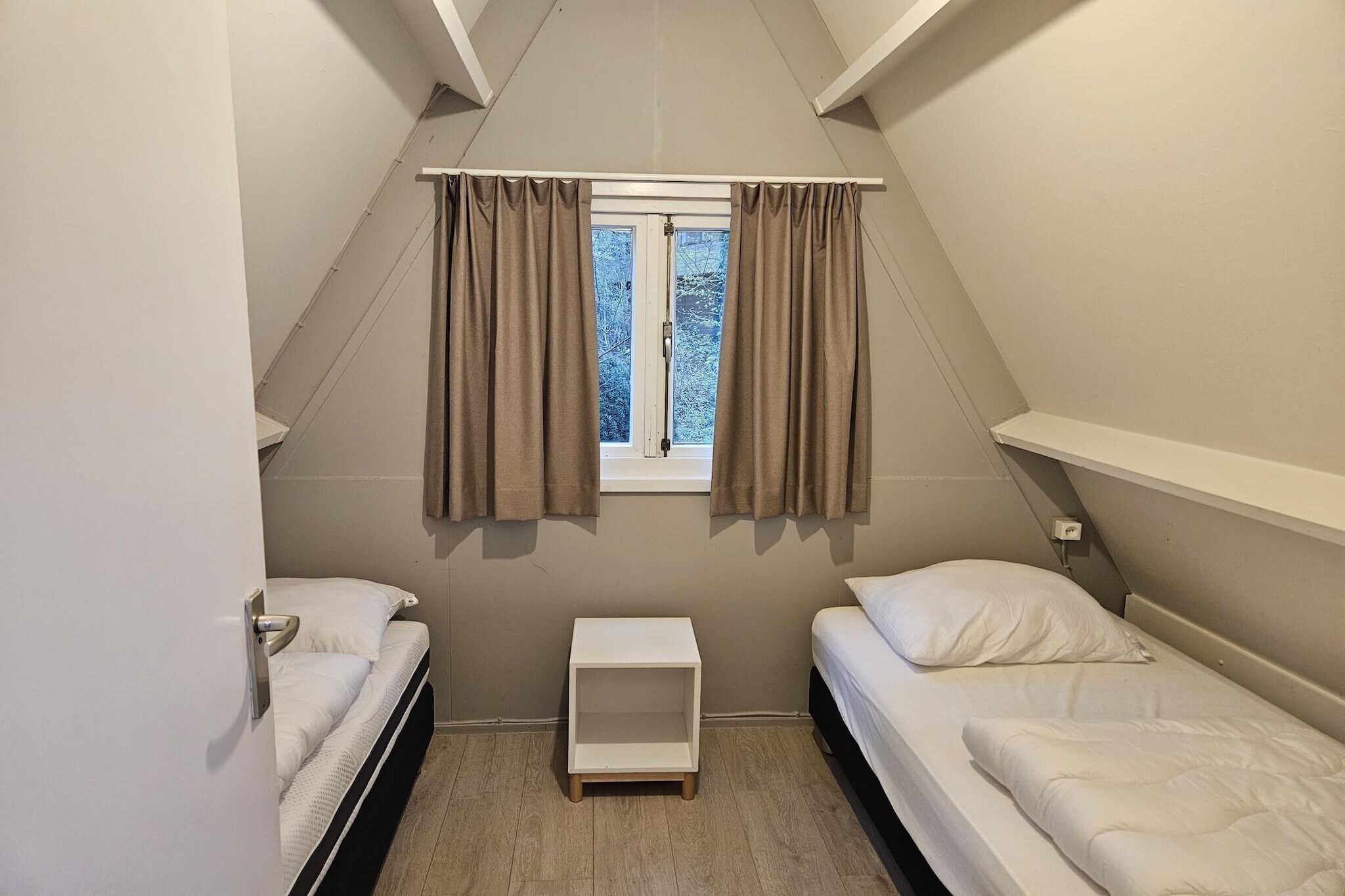 A small, triangular room with two single beds, a window with curtains, and a small white nightstand.