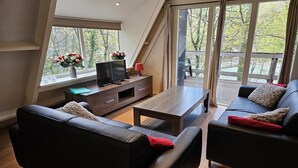A modern living room with a TV, sofa, and coffee table, large windows with a view of trees, and a wooden deck outside. - Serene Vacation Chalet in Durbuy (Durbuy)