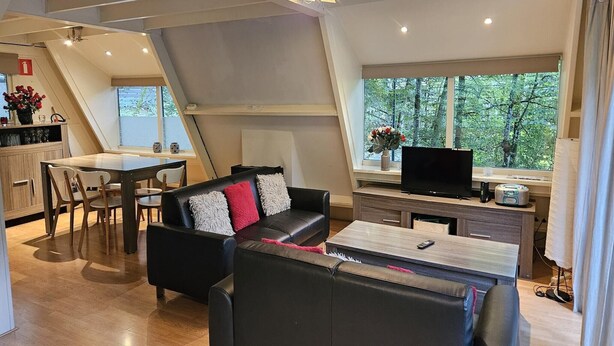 A modern living space with a dining area, a sofa, a TV, and a window with a view of trees. - Serene Vacation Chalet in Durbuy (Durbuy)