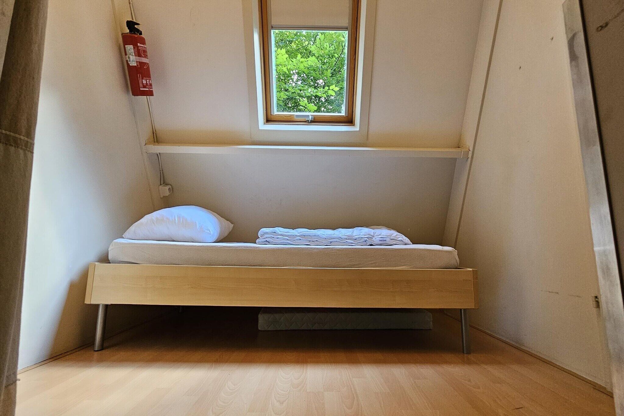 A small, single-bed room with a wooden frame, a window with a view of greenery, and a fire extinguisher mounted on the wall.