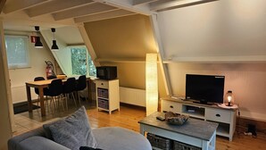A cozy living room with a grey sofa, a TV, and a lamp. There's a dining area with a table and chairs, and a kitchen area with a microwave and a kettle. - Nature Retreat Close to Durbuy Town (Durbuy)