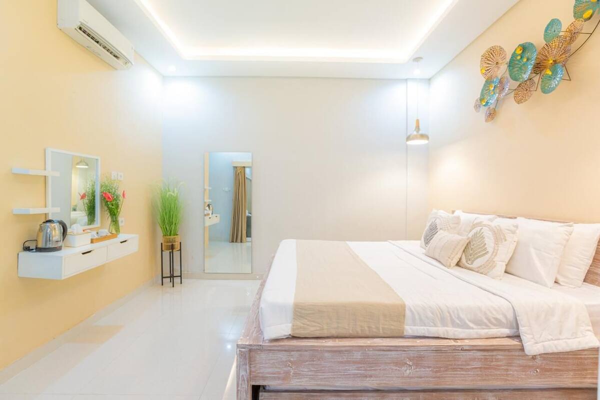 A modern bedroom with a wooden bed, a bedside table with a kettle, a mirror, and a wall-mounted shelf with decorative items.