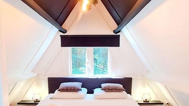 A bedroom with a sloped ceiling, a bed with two pillows, a window with a view of greenery, and a hanging light fixture. - Tranquil Countryside Stay in Durbuy (Durbuy)