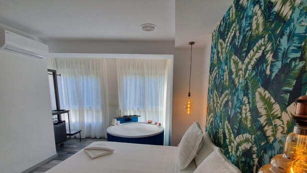 Ein Hotelzimmer mit einem großen Bett, einem kleinen runden Tisch, einer Badewanne und einem großen Fenster mit transparenten Vorhängen.