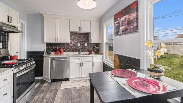 Una cocina moderna con armarios blancos, una estufa negra y un lavavajillas de acero inoxidable. Hay un comedor con una mesa para dos personas, con platos rosas y un florero con flores.