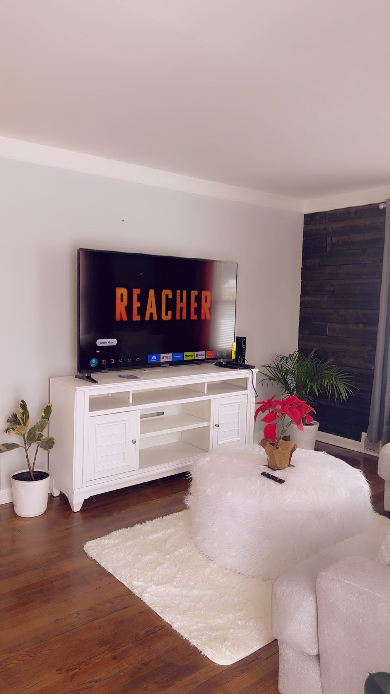 A living room with a flat-screen TV displaying 'REACHER', a white entertainment unit, a fluffy white ottoman, and potted plants.