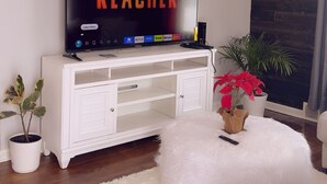 Una sala de estar con un televisor de pantalla plana mostrando 'REACHER', un mueble blanco para equipo multimedia, un otoman blanco mullido y plantas en macetas.